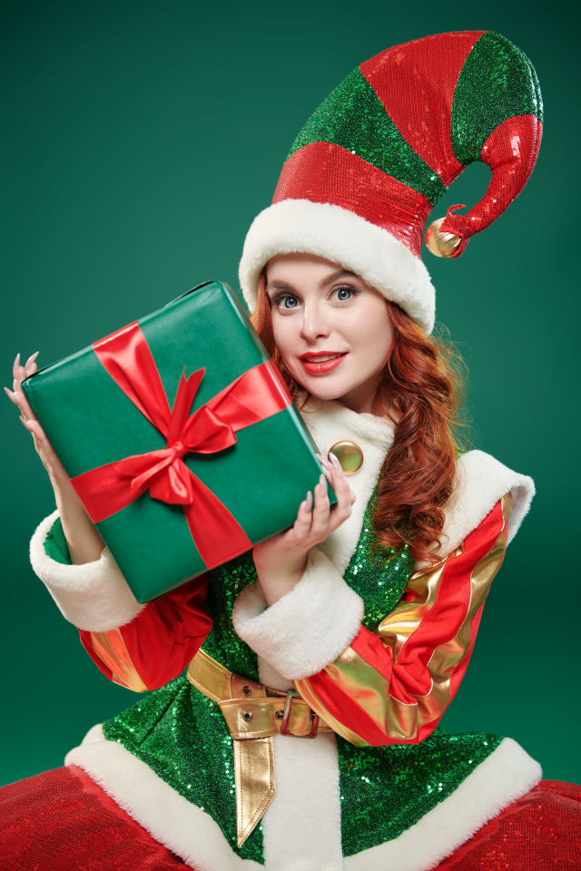 A young redheaded woman in a cute elf outfit, posing with a present against a green background.