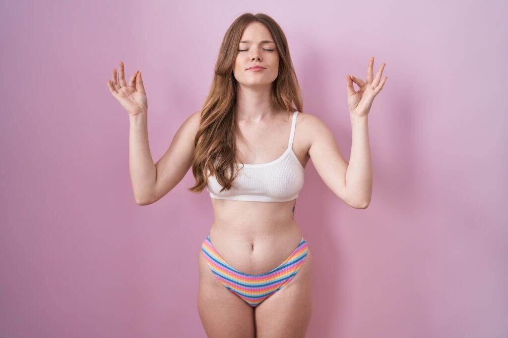 Young white girl standing in her underwear in front of a pink background. She looks zen.