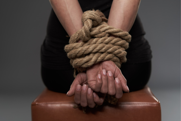 Female sitting on a bench with her hands behind her, bound in thick ropes.