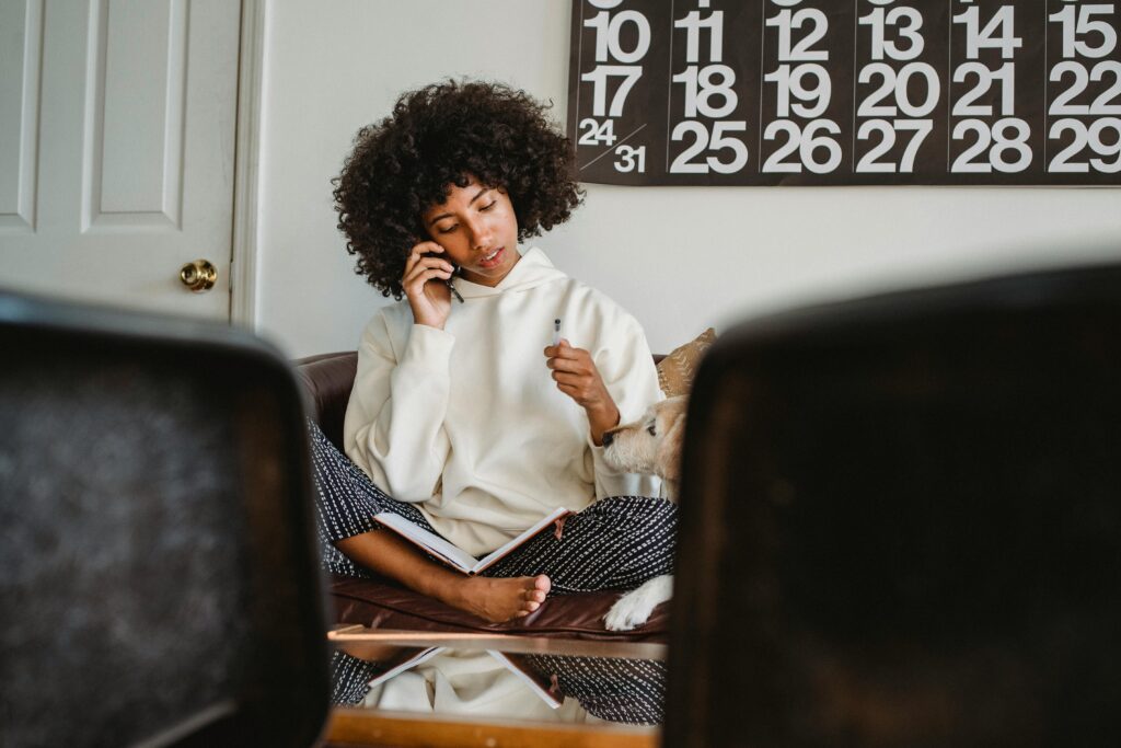 A woman with afro hair is sitting cross-legged on her couch. Talking on the phone.