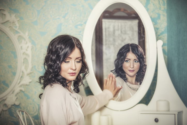 A woman looks away from her reflection in the mirror while her reflection stares at her knowingly.