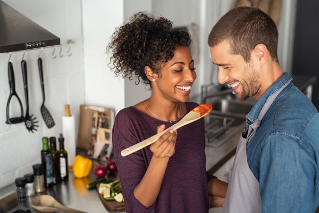 A diverse couple are cooking together in the kitchen. The female is feeding the male a spoon of sauce. They are both smiling.