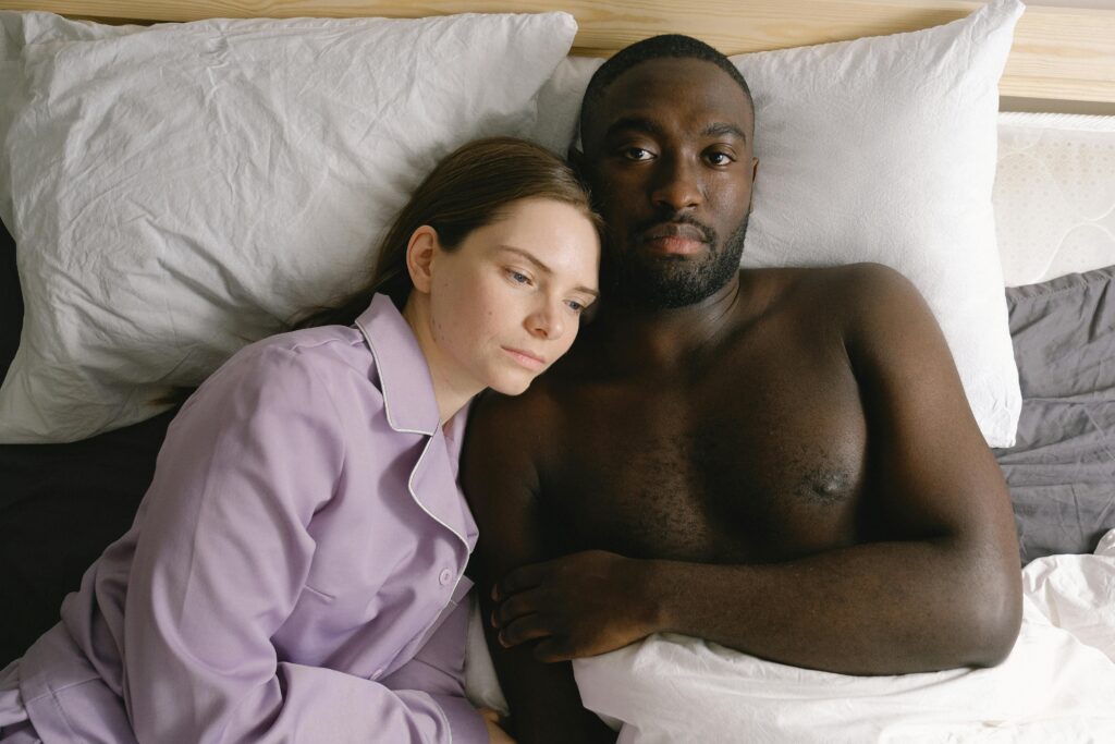A white female and black male couple lay in bed together, both looking unhappy. Possibly due to sexual performance anxiety.