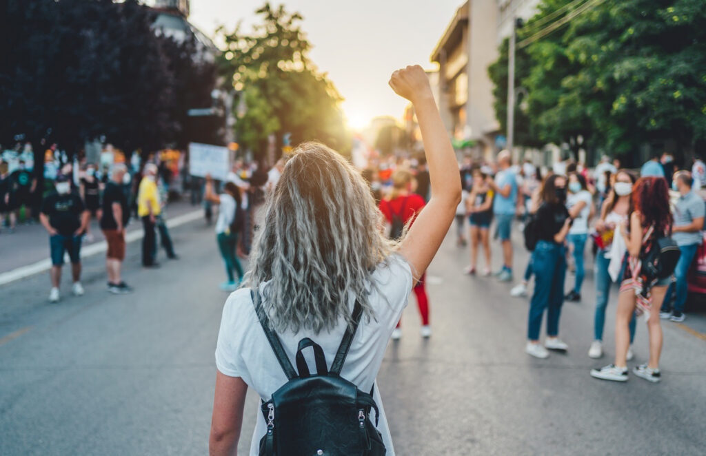A young woman, with her back facing the viewer, her hand in the air making a fist. She stands in front of a group of young protesters on the street.