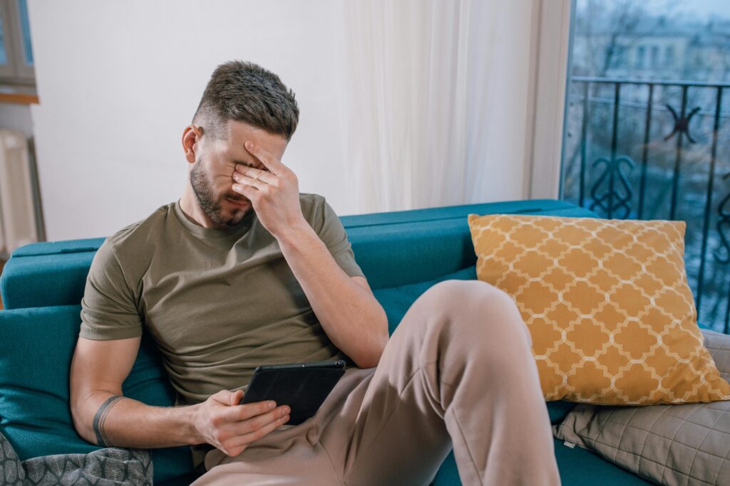 A man sits on a couch with his hand rubbing his eyes. He looks stressed.