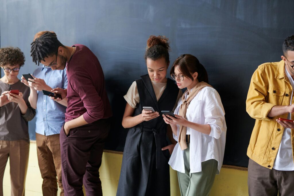 A group of young adults standing in a classroom. They are separated into groups, looking at cellphones. This could be a tech-savvy sex education class.