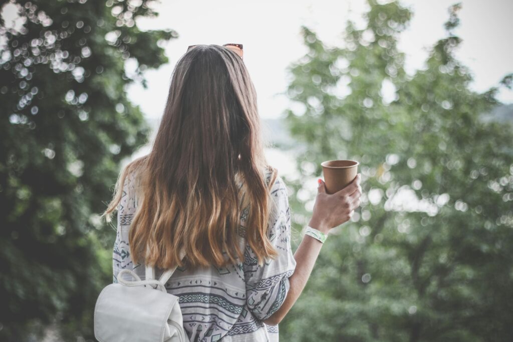 A woman stands with her back turned looking at peaceful scenery while holding a cup.