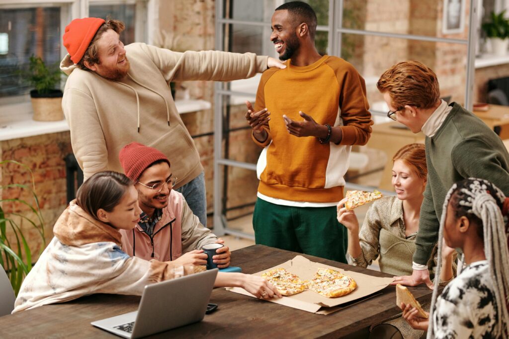 multiracial group of young people surrounding a table, eating pizza together and laughing.